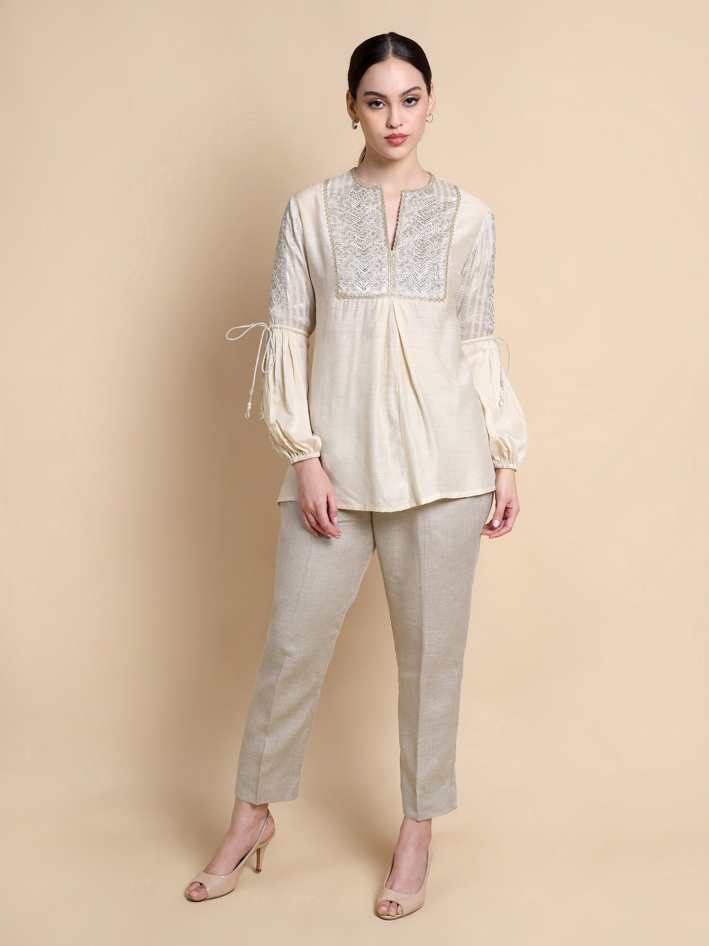 Woman wearing a cream-colored blouse with lace details and beige pants against a beige background