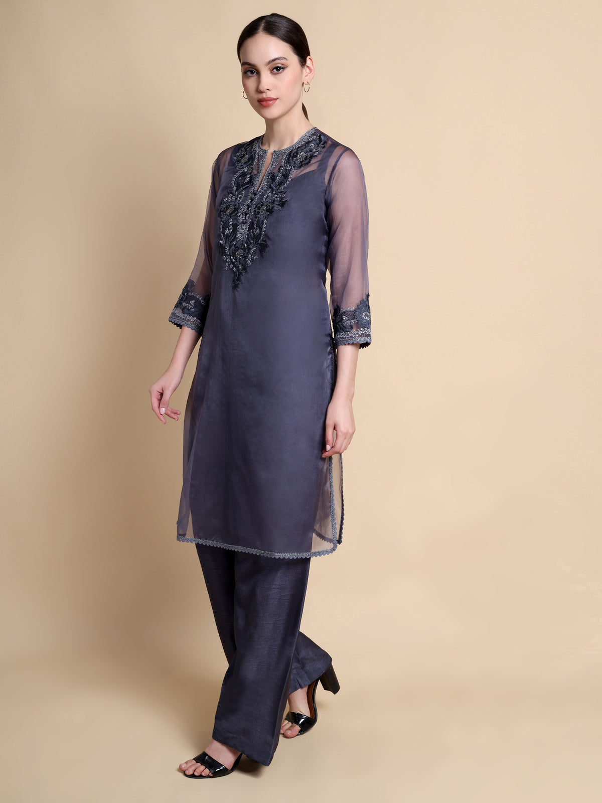 Woman wearing a dark gray traditional outfit with intricate embroidery on a beige background