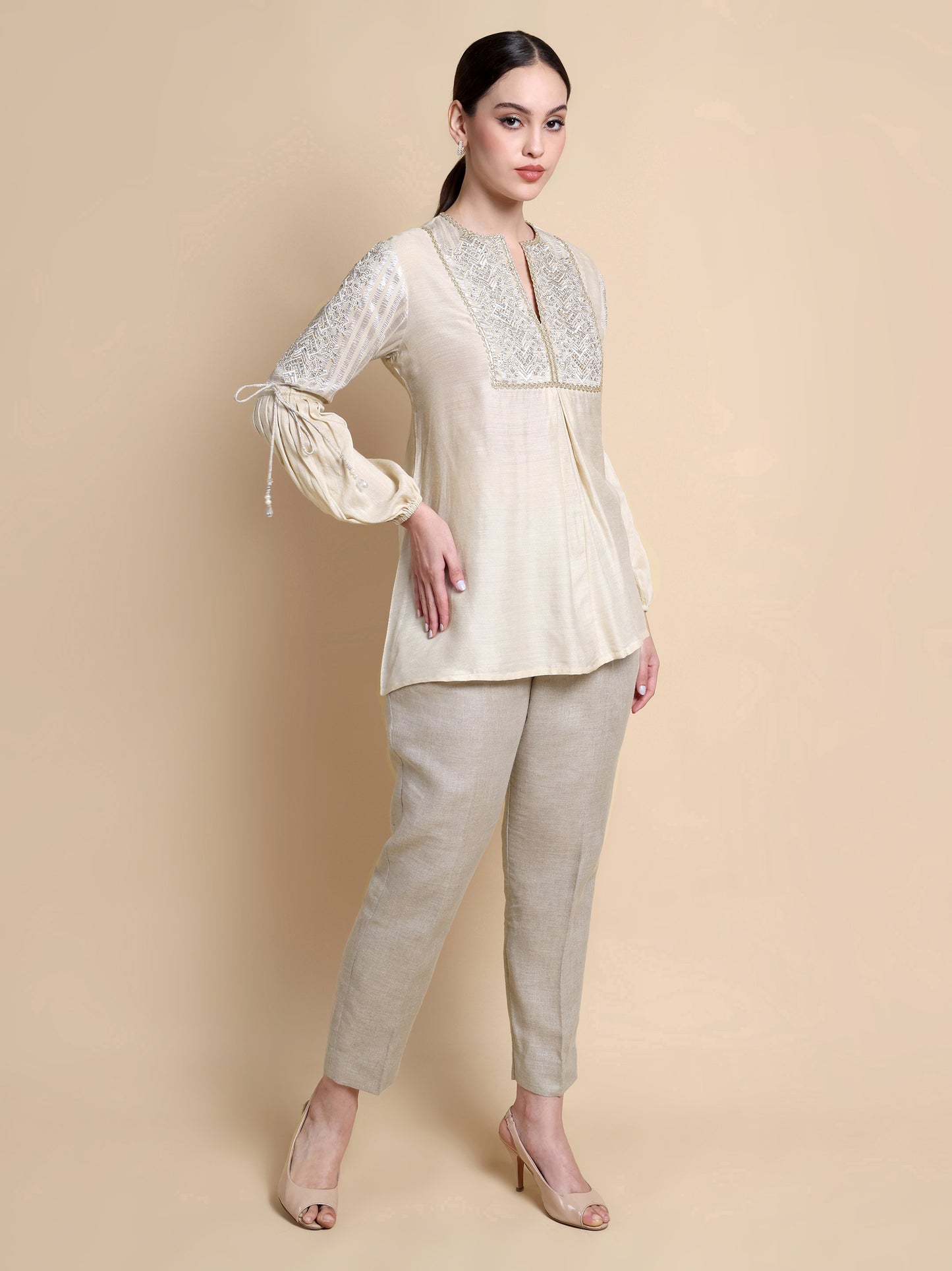 Woman wearing a cream-colored top with lace details and beige pants on a beige background